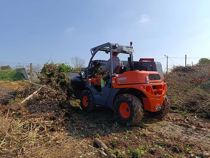 Le chariot télescopique Ausa T144H en action sur un terrain boueux, manipulant des déchets végétaux